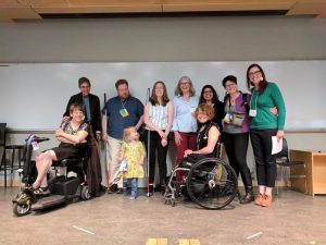 A group of people on a stage, some in scooters, some in wheelchairs, and some standing. Photo of speakers and attendees at the Pacific Western Disability Studies Conference May 2019